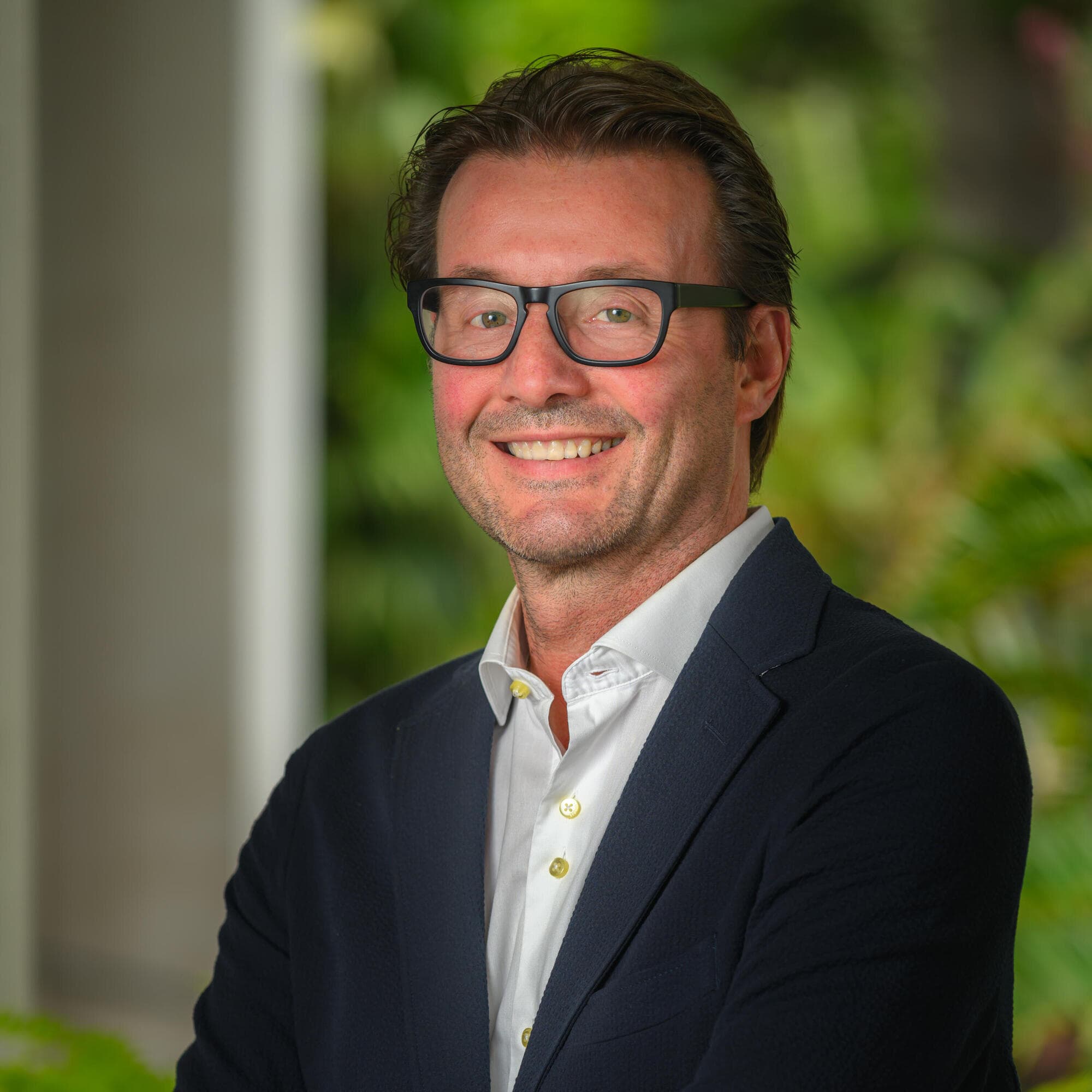 A person in a pale blue button down, dark blue blazer and glasses smiles for the camera.