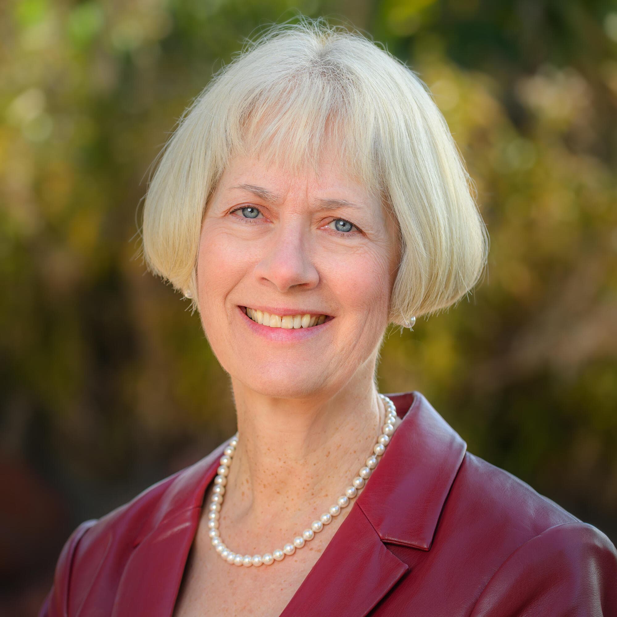 Person with short hair in a red blazer and necklace smiling outdoors.