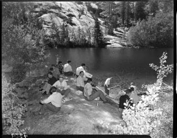 15 men eating lunch and drinking beer on a rock beside a lake in the Big Creek area