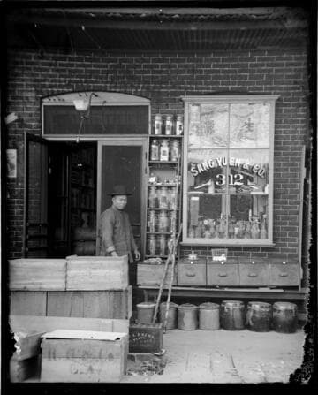 Man standing in front of store, Sang Yuen & Co., with bottled goods in window
