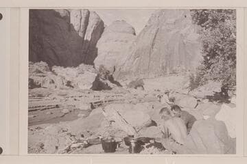 Camp in Navajo Canyon at Pothole Bridge.  Ballard Atherton helps Tom Daly at the kitchen.  Bahe sits in the background.  The creek cuts through left center