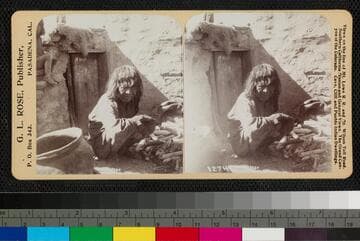 A Hopi woman is crouched near a pile of corn and a pottery bowl, in an unidentified village