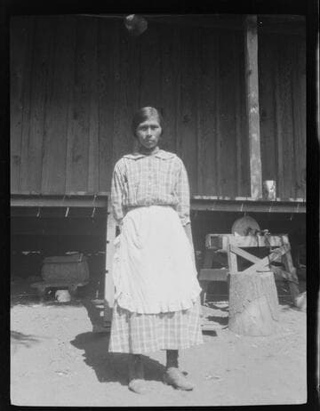 An unidentified Native American woman standing in front of wooden house