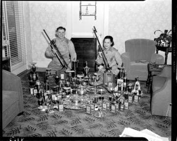 Man and woman holding their competition rifles surrounded by awards and trophys for marksmanship