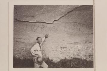 J. W. Black name with date Feb 2, 1894 written with charcoal in a cave on the right bank above the Escalante River. Harry Aleson stands at the wall