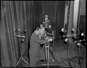 Cinematographer filming two ladies seated on stage. Man holding slate in back of them says: "Its So Easy"