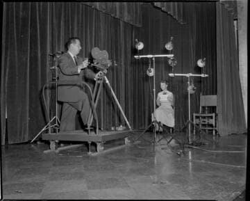 Cinematographer with 16mm movie camera on dolly filming lady seated on stage of Edison auditorium. She's holding a card with #23 on it